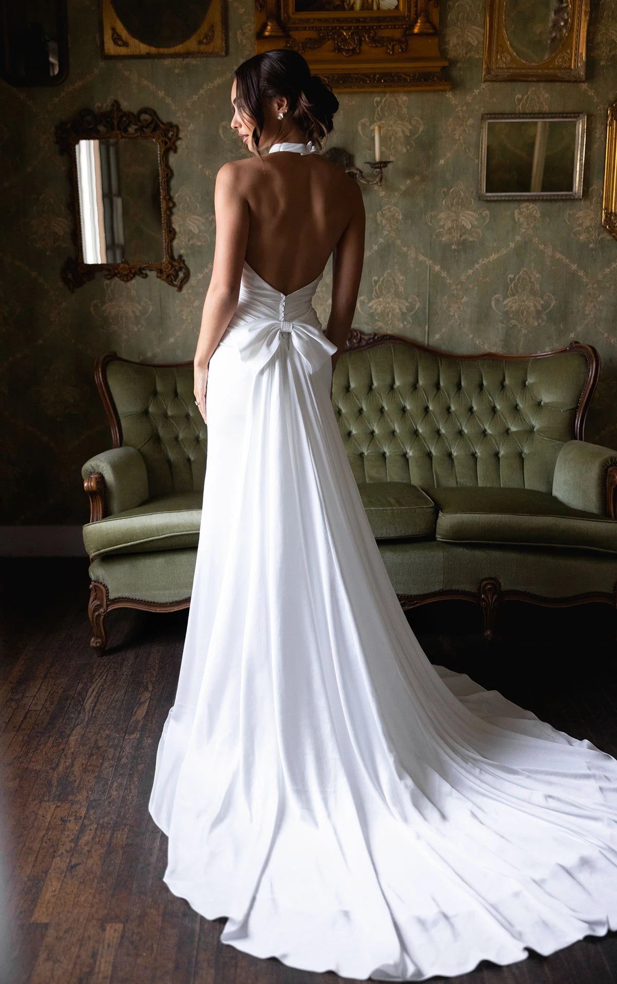 Woman in a white gown with a bow detail standing in an elegant room with green sofas and mirrors.