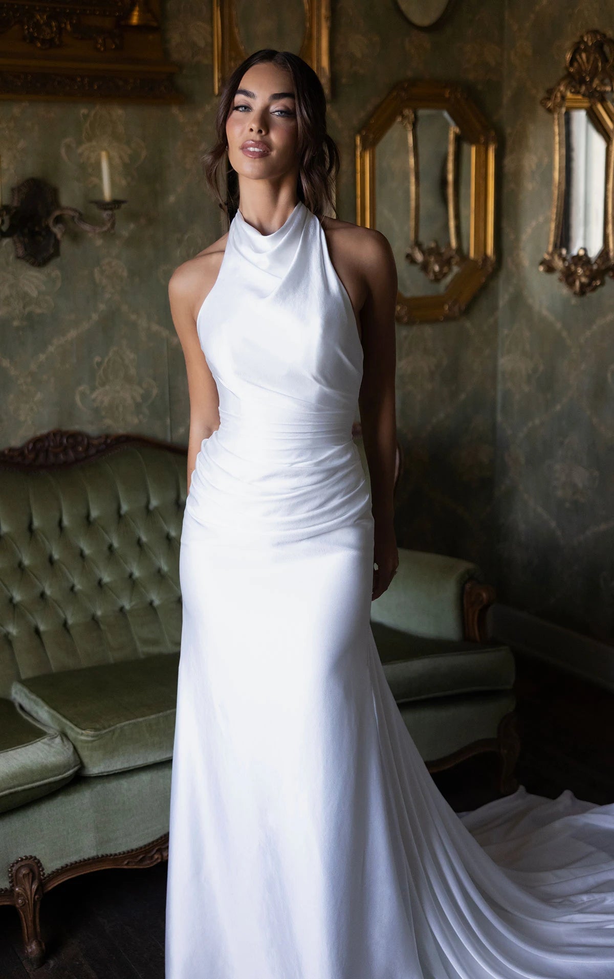 Woman in a white dress standing in an ornate room with mirrors and a couch.